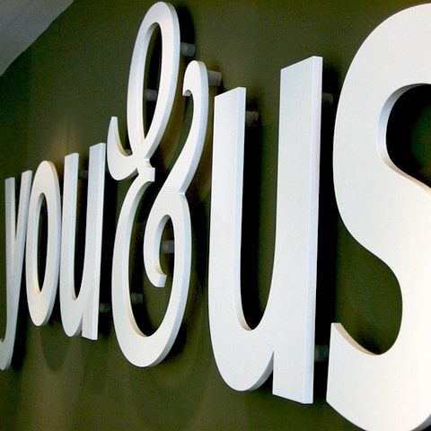You&Us white logo made from flat cut letters mounted to olive green wall by Fabricut